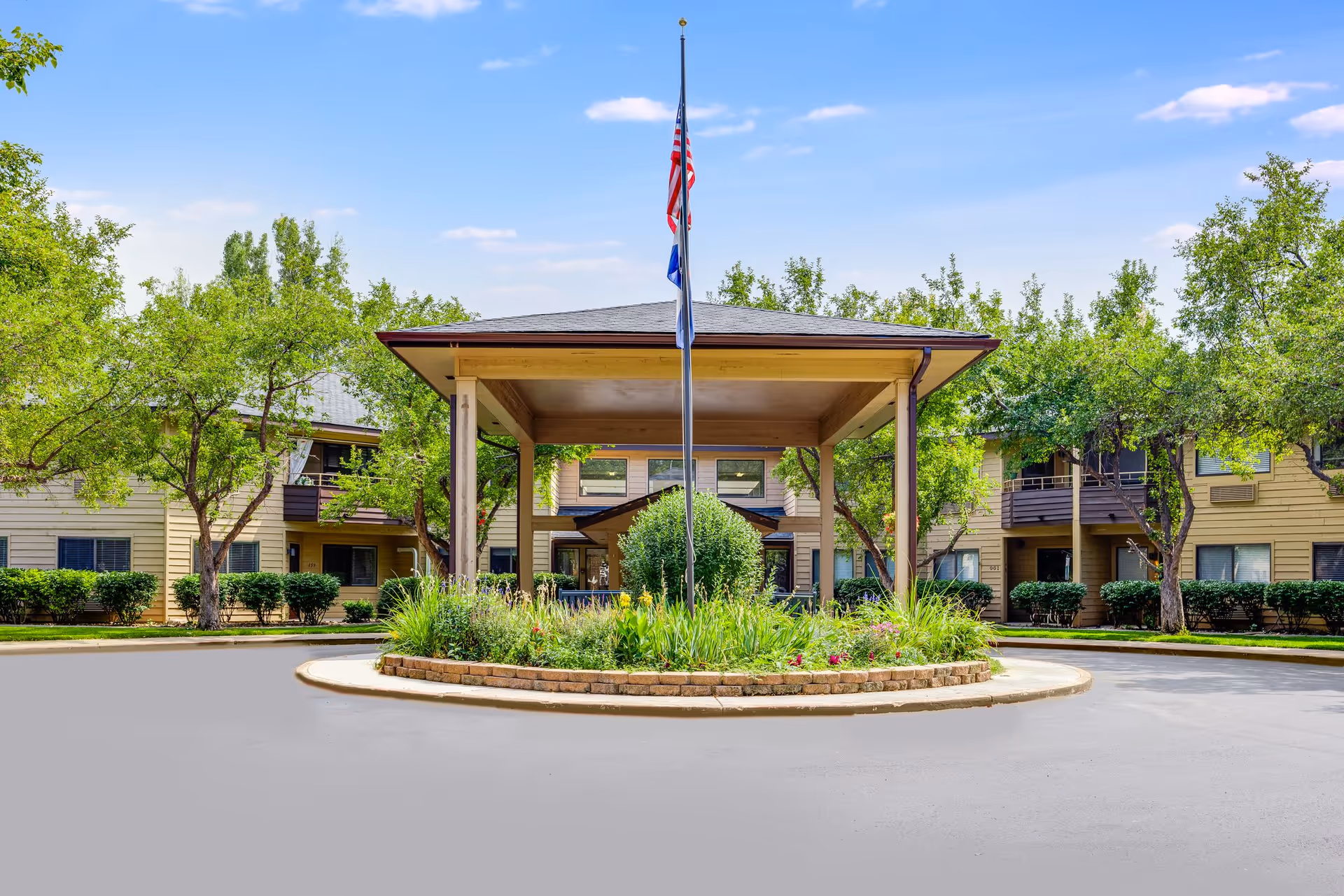 Covered entrance with a flagpole and landscaped roundabout in front of a two-story senior living building surrounded by trees.