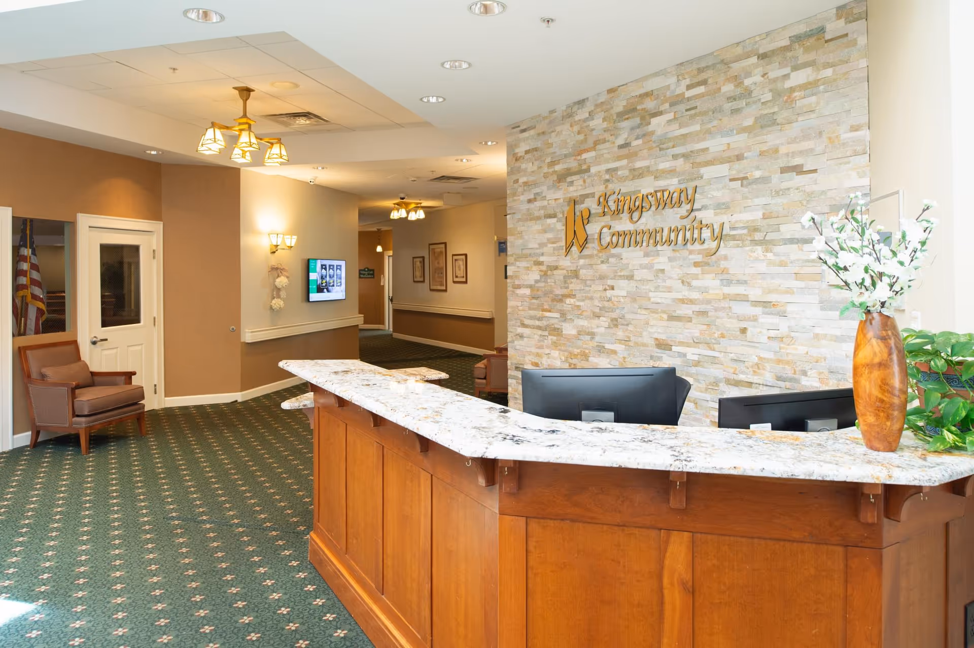 Bright reception lobby with a marble-topped wooden front desk, stone accent wall reading 'Kingsway Community', seating, and a vase of flowers.