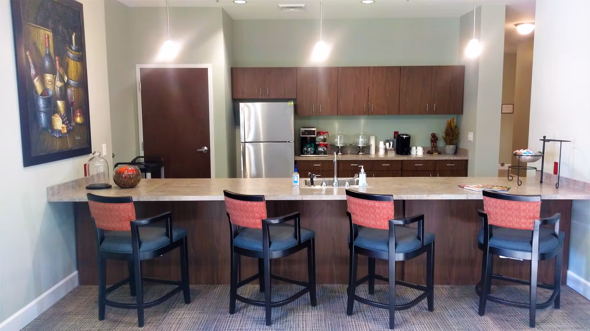Communal kitchenette with a long countertop, four bar stools, stainless refrigerator, upper cabinets and a coffee station.