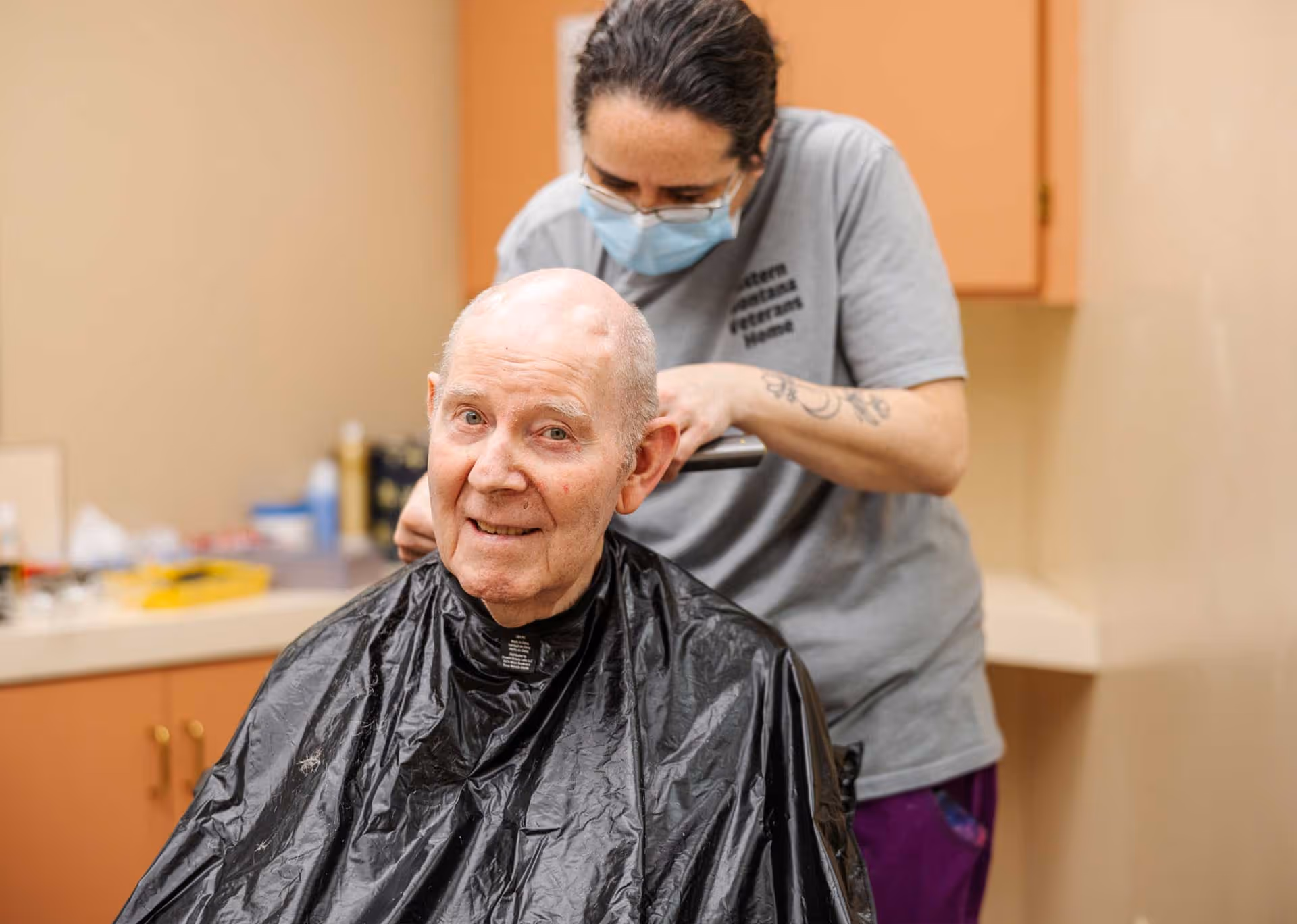 An elderly man wearing a black haircut cape is getting a haircut from a caregiver wearing a face mask and a gray shirt with the text Eastern Montana Veterans Home in a room with beige walls and wooden cabinets.