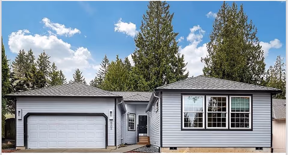Front exterior of a gray single-story house with an attached two-car garage, large windows, and tall trees behind under a blue sky.