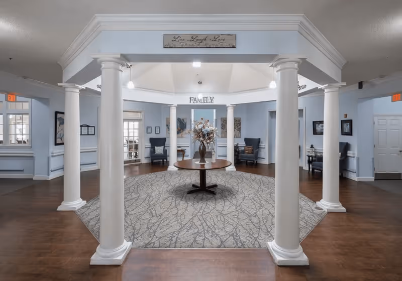 Interior view of a senior living facility lobby area with white columns surrounding a round table with a floral arrangement. The walls are painted light blue and decorated with framed artwork. There are several chairs placed around the room and signs that read 'Live Laugh Love' and 'Family'. The floor is a combination of wood and a patterned carpet.