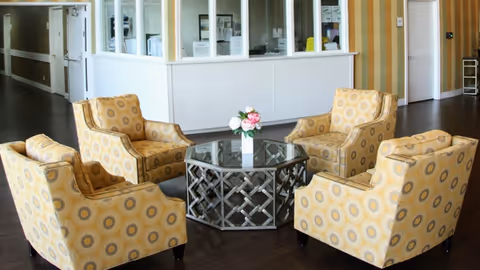 Four yellow patterned armchairs arranged around a glass-topped coffee table with a small floral centerpiece in a facility lobby.