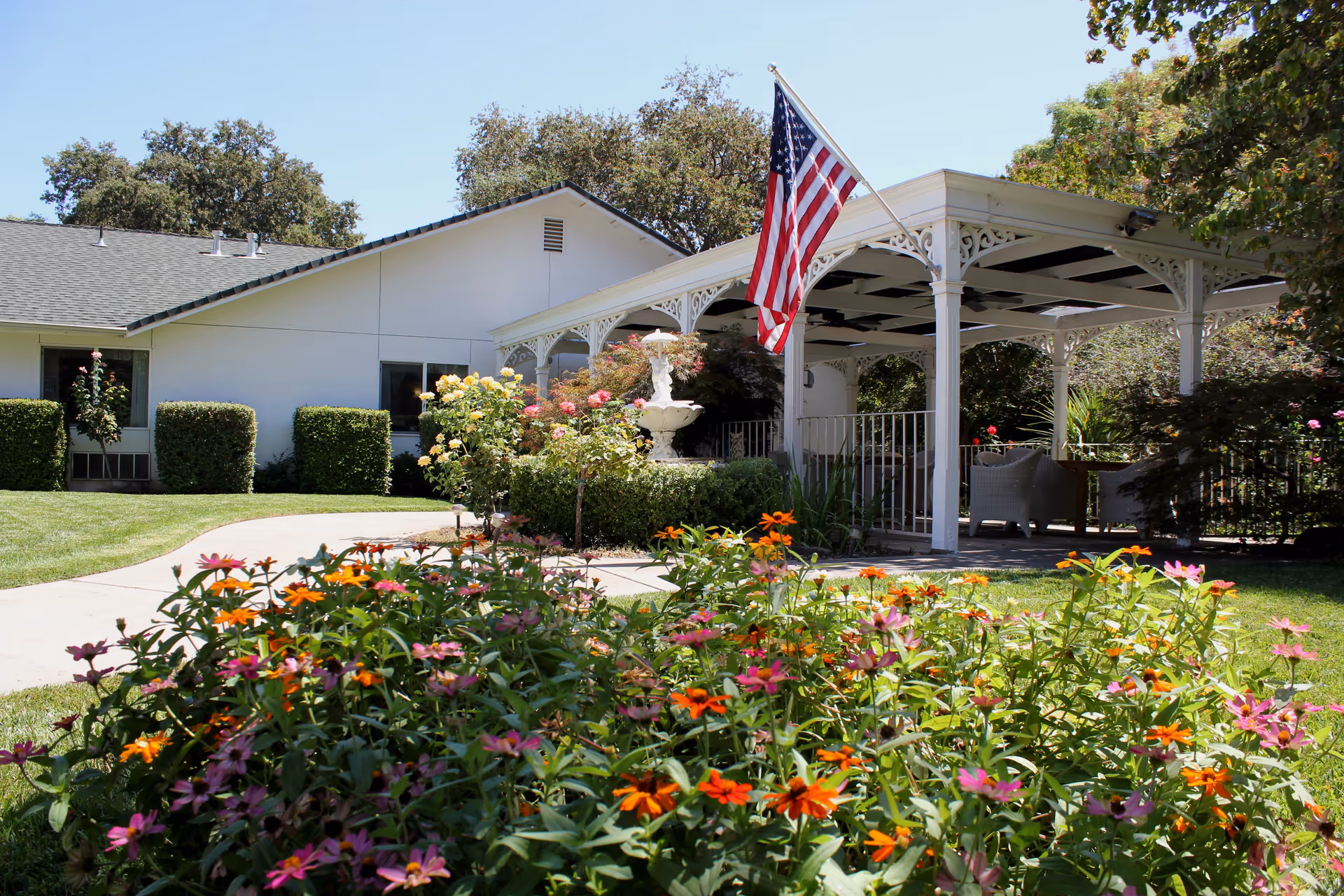 Outdoor garden area at Roses & Ivy Assisted Living Facility for the Elderly featuring a variety of colorful flowers in the foreground, a white gazebo with decorative trim and seating underneath, an American flag mounted on the gazebo, a white fountain surrounded by bushes, and a white building with a sloped roof in the background under a clear blue sky.