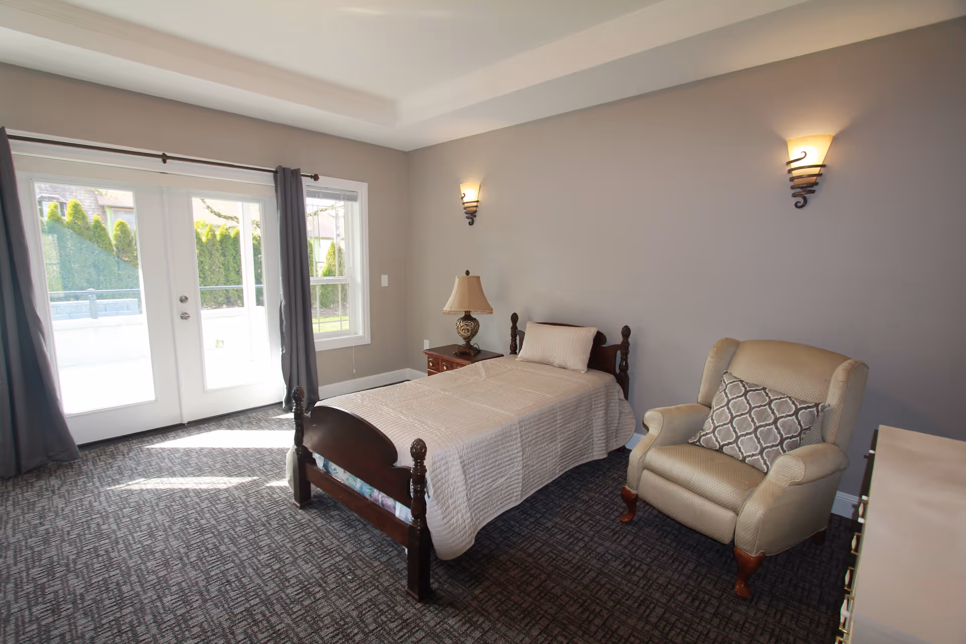 A bright and clean bedroom with a single wooden bed covered with a beige quilt and a pillow. Next to the bed is a wooden nightstand with a decorative lamp. There is a comfortable beige armchair with a patterned cushion on it. The room has gray walls with two wall-mounted lights and a large window and glass door letting in natural light, showing a view of greenery outside.