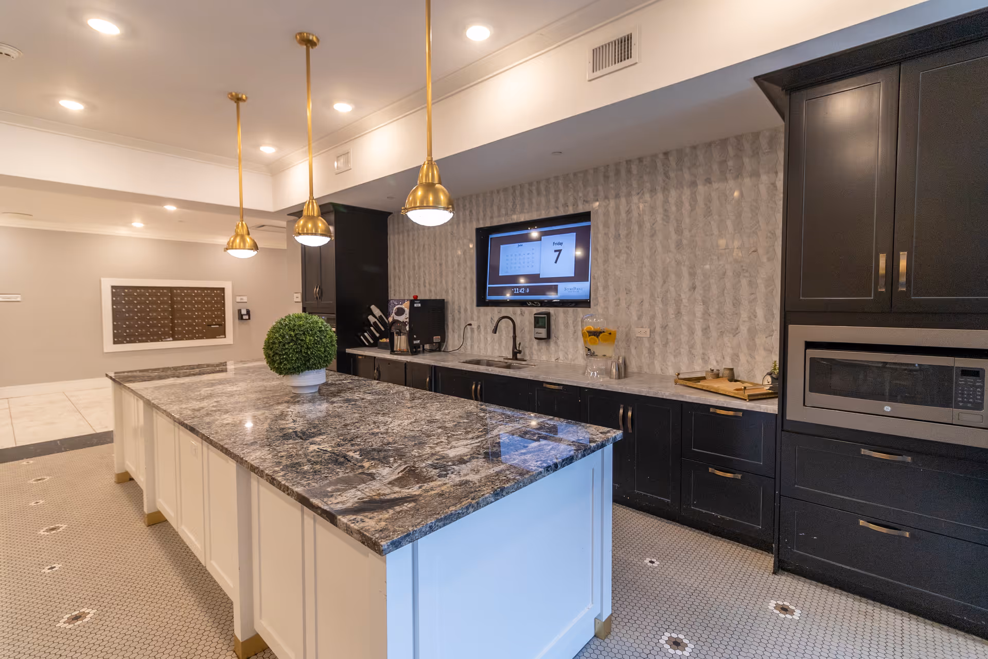 Modern kitchen area with a large marble island countertop, white cabinetry, and three gold pendant lights hanging from the ceiling. The back wall features black cabinets, a microwave, a sink, a coffee machine, and a mounted digital screen displaying the date and time. The floor has a hexagonal tile pattern with small flower-like designs.