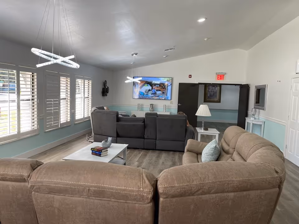 A bright and spacious living room area with large windows featuring white shutters on the left wall. The room contains two large sectional sofas, one beige and one gray, arranged around a white coffee table with books and a decorative bird sculpture. A flat-screen TV is mounted on the far wall above a small console table. Modern circular pendant lights hang from the ceiling, and there is a side table with a lamp next to the beige sofa. The walls are painted white with a light blue wainscoting, and the floor is a light wood laminate.