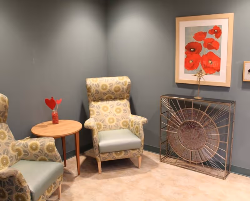A cozy corner with two patterned armchairs featuring light green cushions, a small round wooden table between them with a red vase holding heart-shaped decorations, and a decorative metal and glass console table against a gray wall. Above the console table hangs a framed artwork of red poppies.