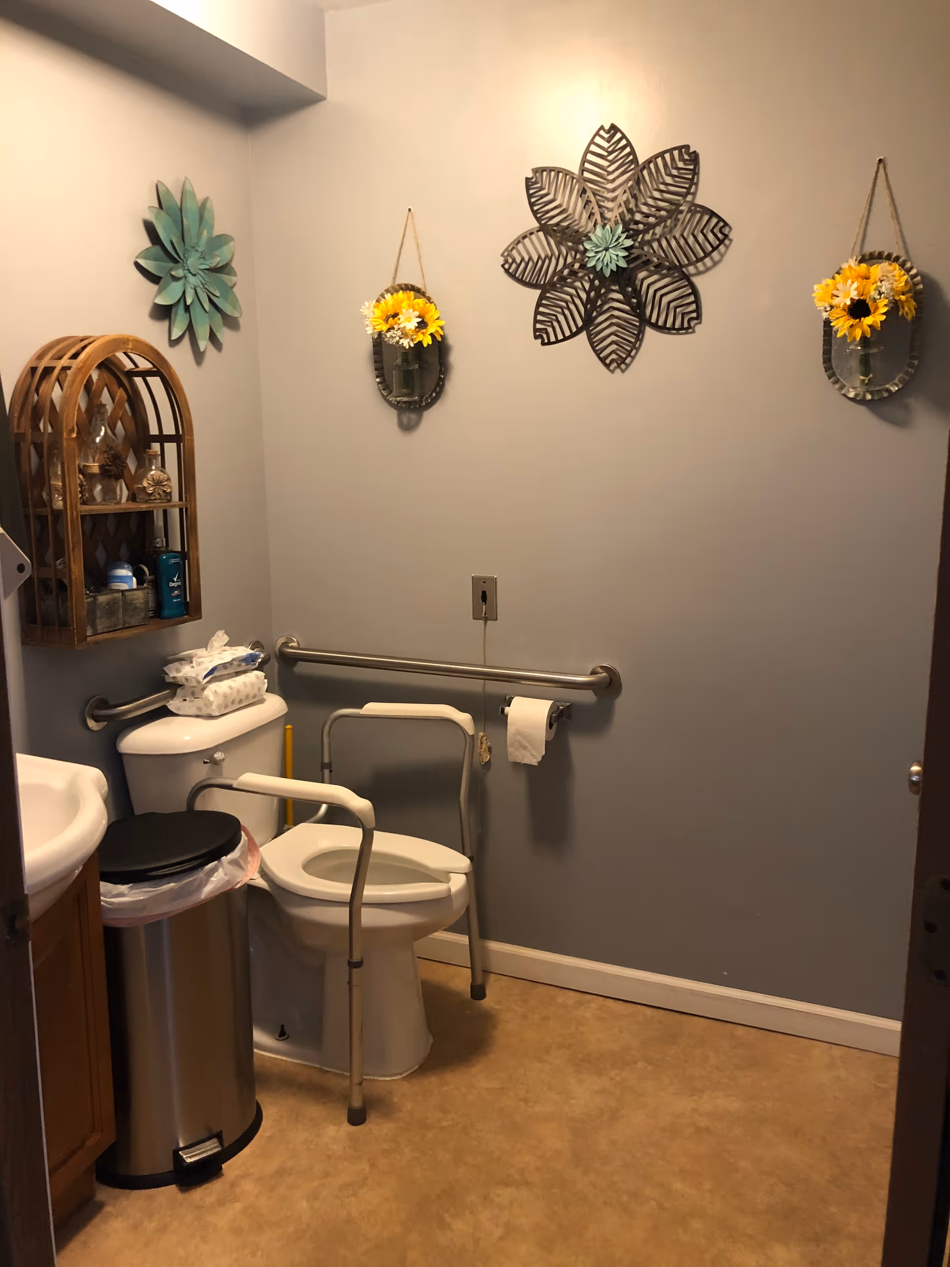 A small bathroom with a toilet equipped with safety rails, a trash can, and a wooden wall shelf holding toiletries. The walls are decorated with three flower-themed wall hangings, including two small metal containers with yellow flowers and a large metal flower in the center.