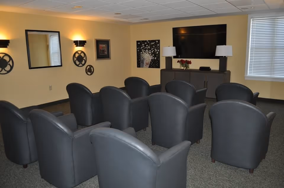 Small theater-style common room with rows of gray armchairs facing a wall-mounted TV and cabinet.