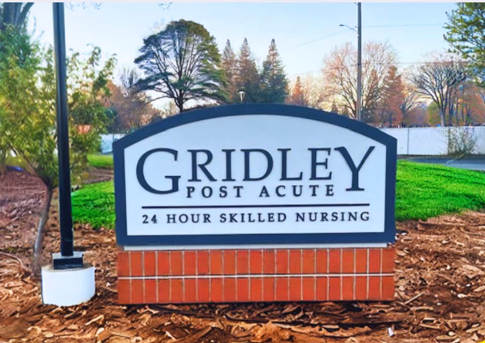 Outdoor sign for Gridley Post Acute, a 24 hour skilled nursing facility, set on a brick base with trees and greenery in the background.
