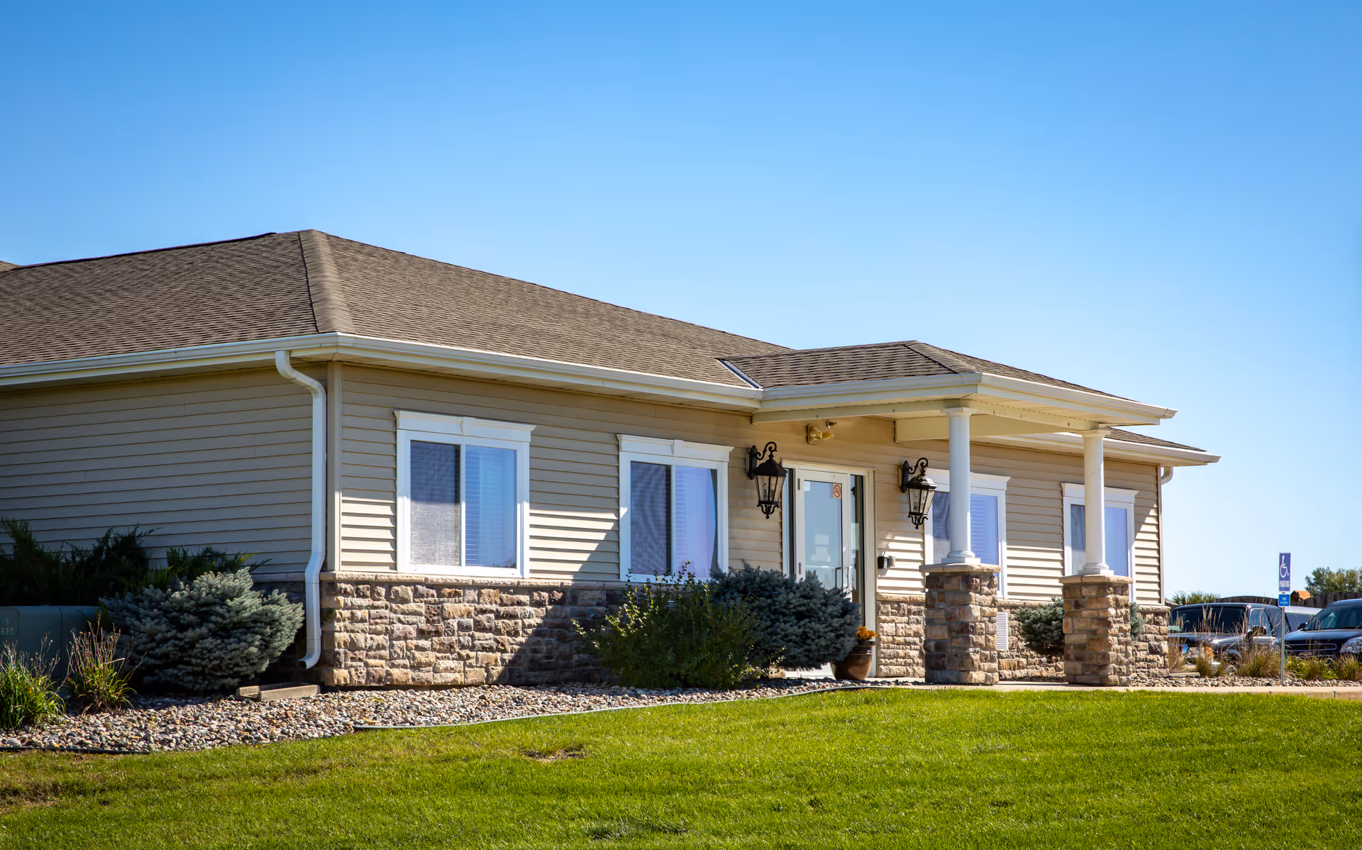 Exterior view of a single-story building with beige siding and stone accents, a covered entrance supported by white columns, surrounded by green grass and landscaping under a clear blue sky.