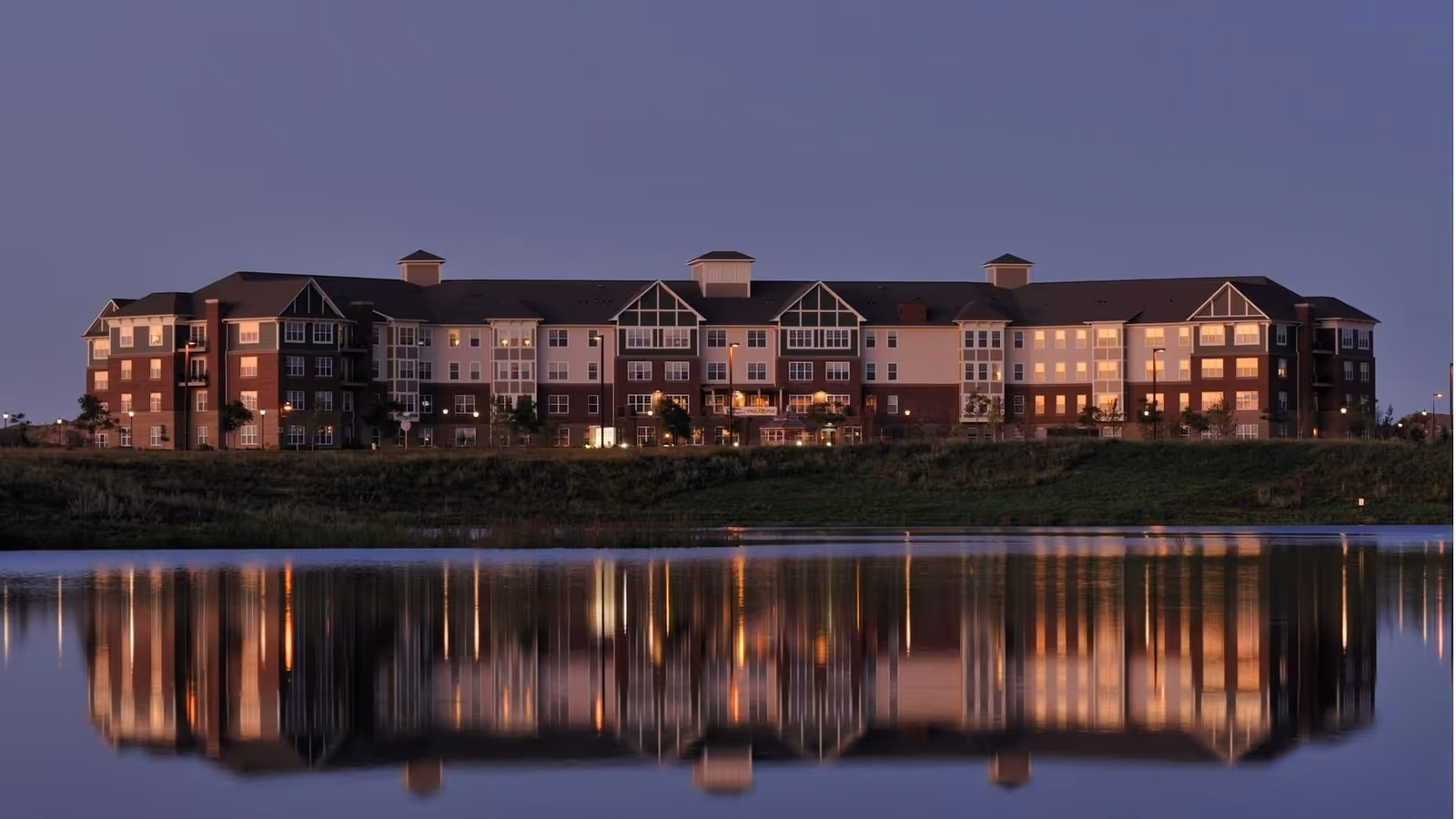 Wide exterior view of a large senior living facility building at dusk with lights on inside, reflected in a calm body of water in the foreground.