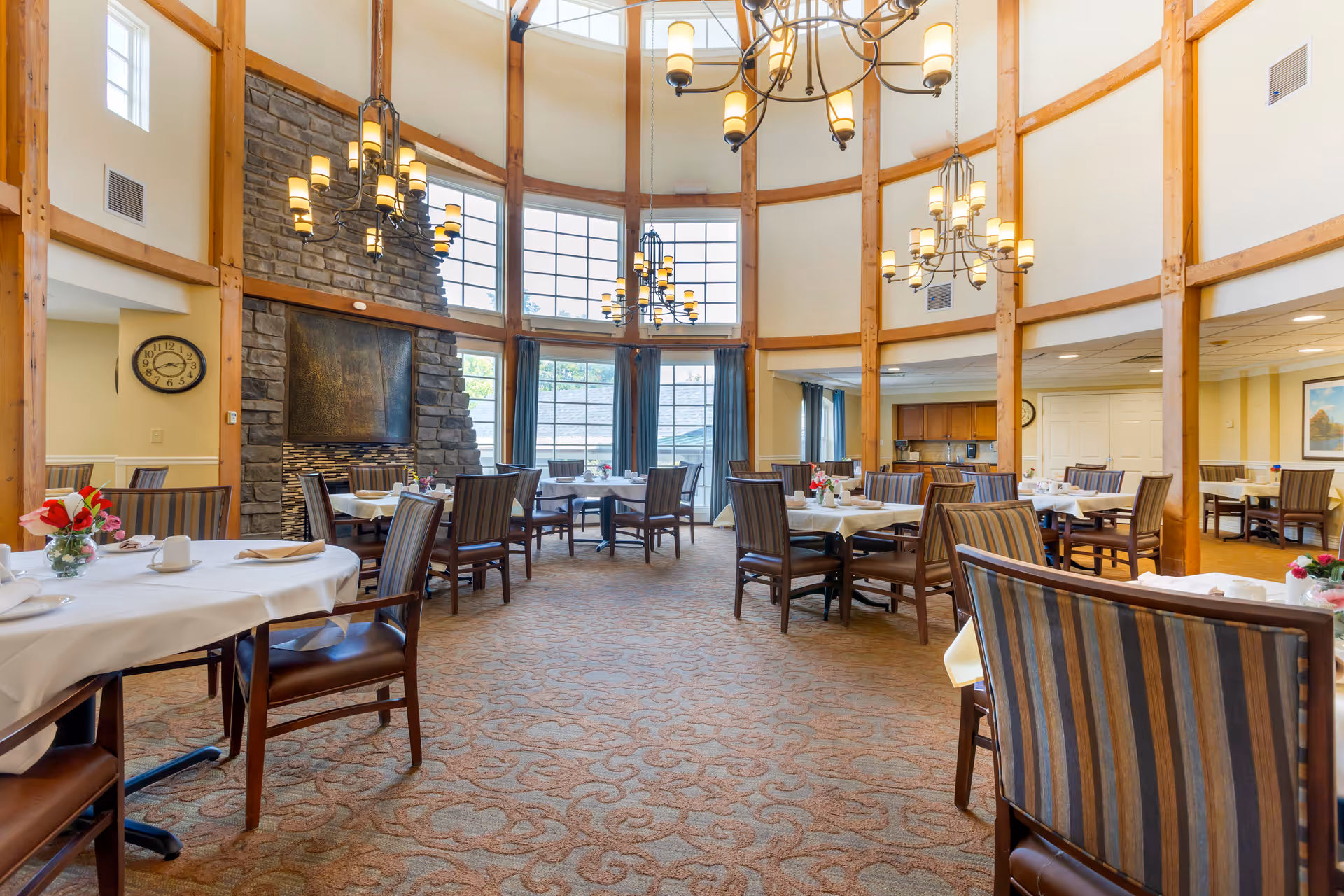 A spacious dining room with multiple tables covered in white tablecloths and set with plates, cups, and napkins. The room features high ceilings with large windows letting in natural light, wooden beams, and several elegant chandeliers. There is a stone fireplace on one side and striped upholstered chairs around the tables. The carpet has a patterned design, and there are floral centerpieces on the tables.