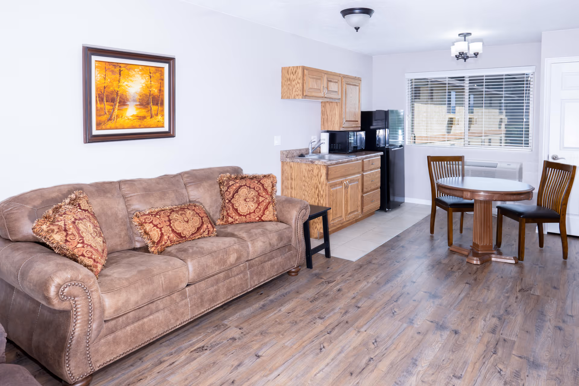 Interior view of a senior living facility room featuring a brown upholstered sofa with three decorative pillows, a small kitchenette with wooden cabinets, a microwave, and a black refrigerator. There is a round wooden dining table with three chairs near a window with blinds. A framed painting of a sunset over a forest hangs on the wall above the sofa.