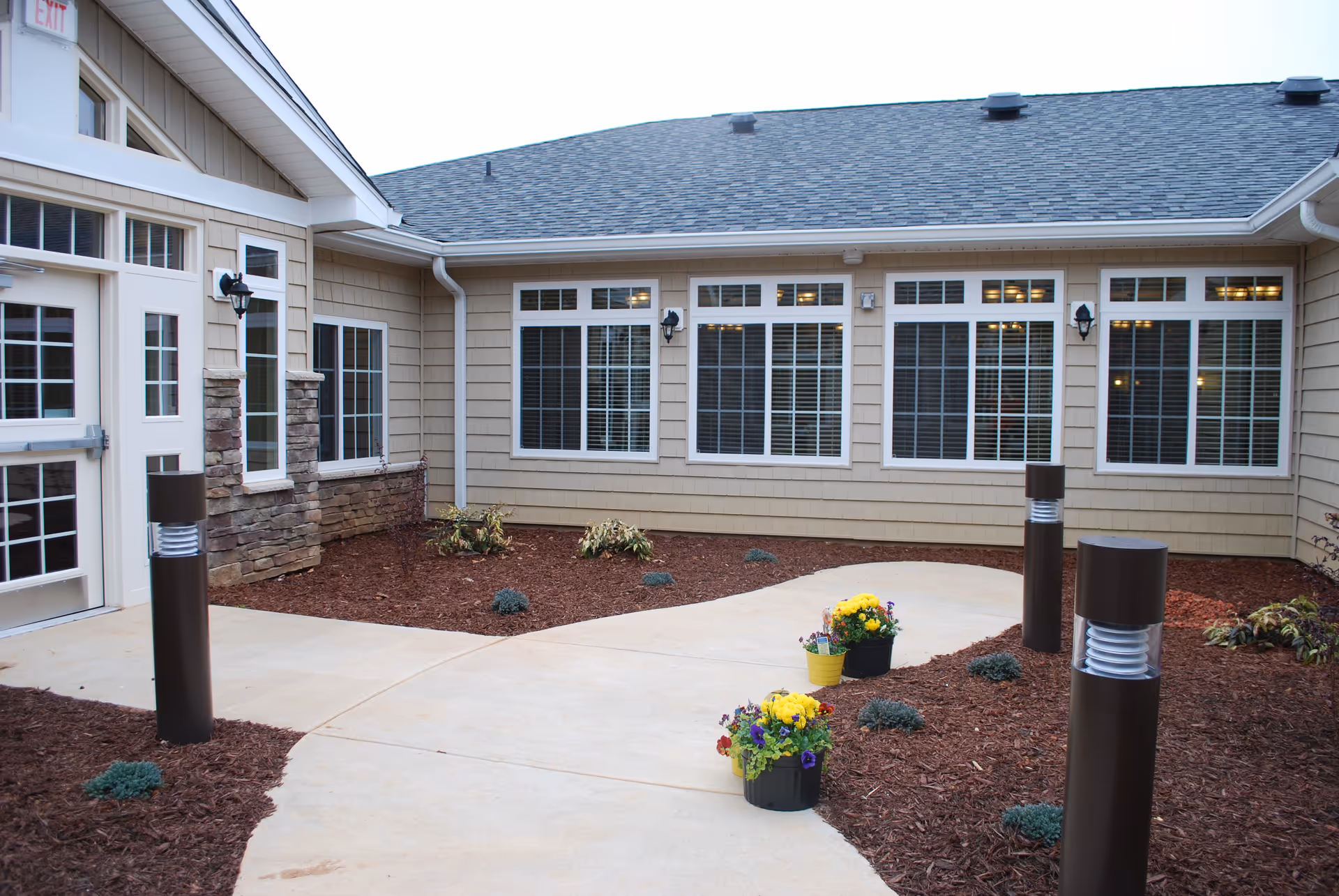 Paved courtyard with a winding walkway, flower pots, and exterior windows of a senior living facility.