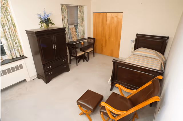 Furnished bedroom with a single wooden bed, dark wood armoire, small table with chairs, and a leather armchair with ottoman.