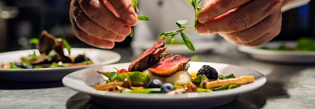 Close-up of a chef's hands garnishing a gourmet dish with fresh herbs, featuring slices of rare-cooked meat, mashed potatoes, berries, and vegetables on a white plate.