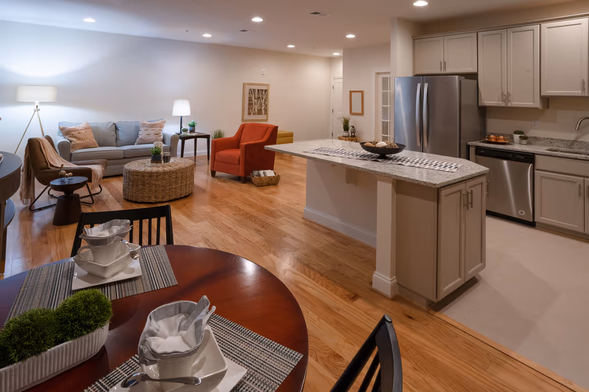 Open concept living area with a kitchen and dining space. The kitchen features light-colored cabinets, a granite countertop island, stainless steel refrigerator and dishwasher. The living room has a gray sofa, an orange armchair, a brown chair, a round wicker coffee table, and two lamps providing warm lighting. The dining table is set with white dishes and napkins, and there are small green plants as decoration.