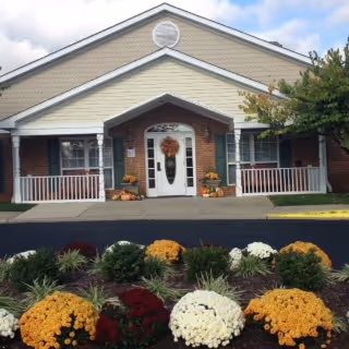 Front exterior view of a single-story building with a peaked roof and a covered entrance decorated with a fall wreath on the door. There are flower pots with autumn-themed plants on either side of the entrance and a landscaped garden with colorful flowers in the foreground.