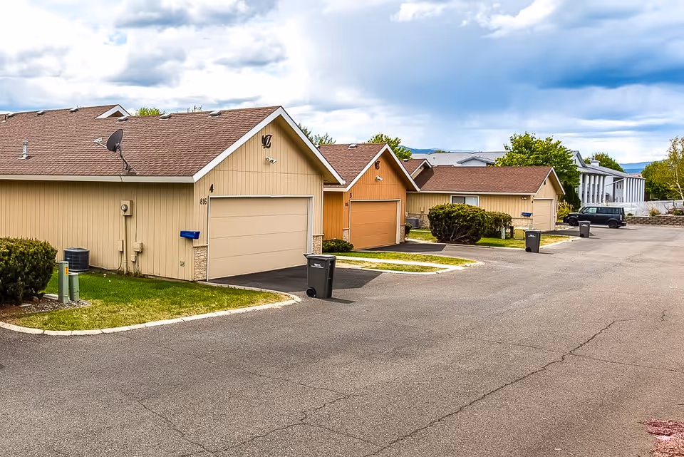 A row of single-story residential buildings with attached garages, beige and light brown siding, and brown shingle roofs. The buildings are situated along a paved driveway with trash bins placed near the garages. There are some bushes and green grass patches around the buildings, and a partly cloudy sky overhead.