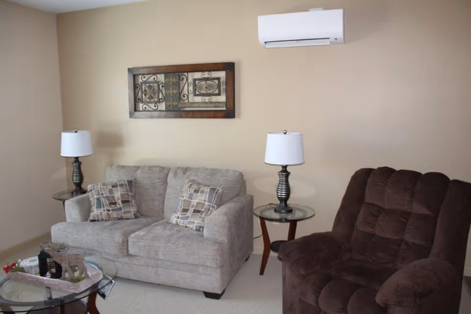 A cozy living room with a beige loveseat adorned with two patterned pillows, a dark brown cushioned recliner chair, two glass-top side tables each with a lamp, a glass coffee table with decorative items, a beige wall with a decorative framed metal wall art, and a white air conditioning unit mounted on the wall.