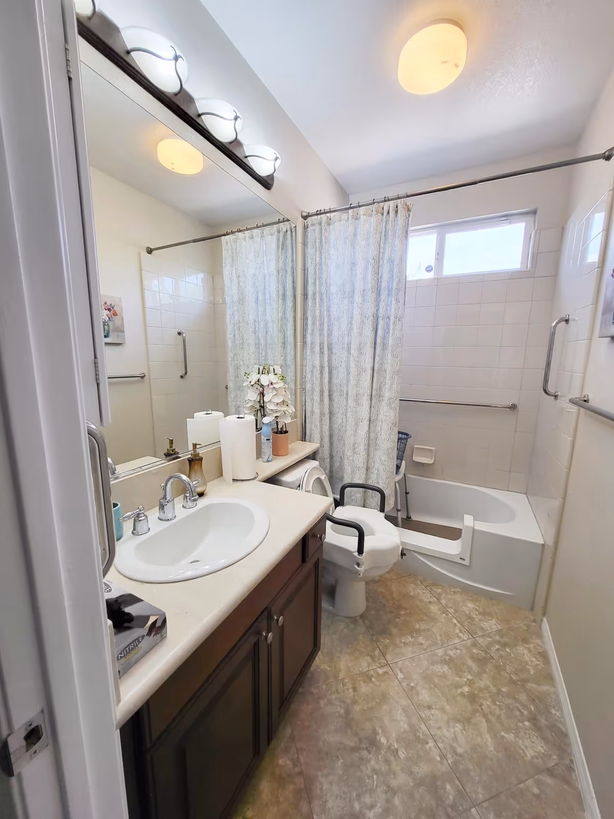 A bathroom with a sink and countertop on the left, a large mirror above the sink, and a toilet with armrests next to a bathtub with a shower curtain. The bathtub has grab bars installed on the walls for safety. The floor is tiled, and there is a window above the bathtub allowing natural light in.