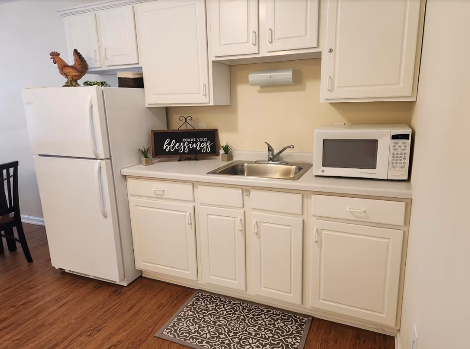 A small kitchen area with white cabinets, a stainless steel sink, a white microwave on the countertop, and a white refrigerator. There is a decorative sign on the counter that reads 'count your blessings' along with two small potted plants. A patterned rug is placed on the wooden floor in front of the sink.