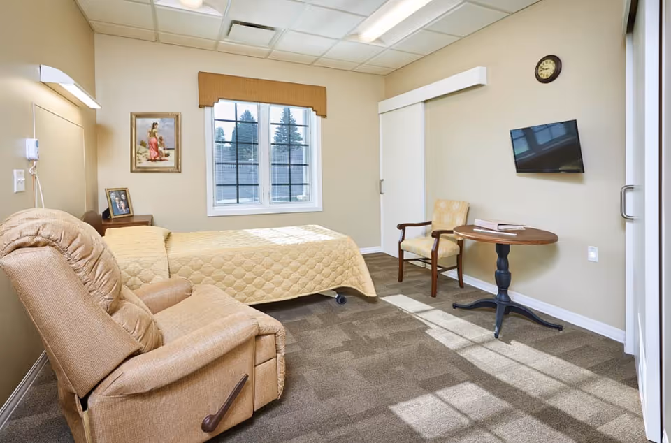 A bright and clean bedroom in a senior living facility featuring a beige recliner chair, a bed with a yellow quilt, a wooden side table with a framed photo, a window with a brown valance, a wall-mounted TV, a round wooden table with a book, and a cushioned armchair. The room has beige walls, carpeted flooring, and a clock on the wall.