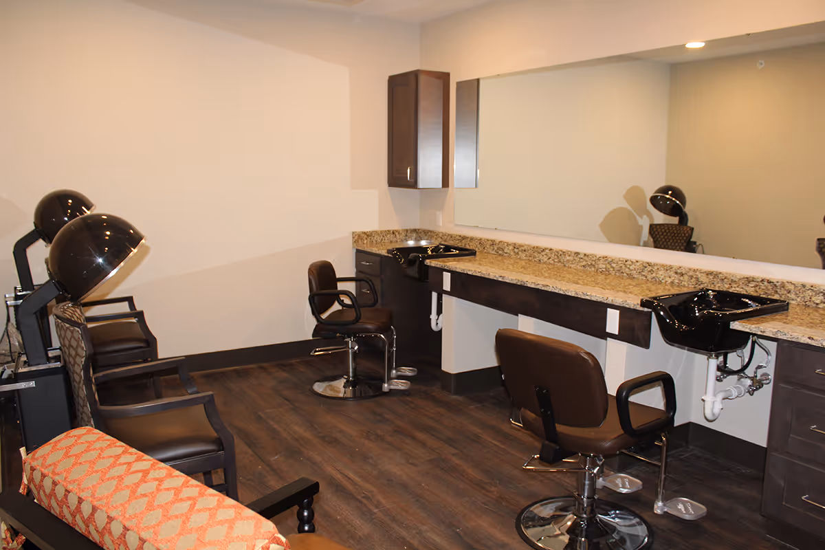 Interior view of a salon area with two black hair washing sinks, two brown salon chairs, two black hair dryer chairs with hooded dryers, a large mirror above a granite countertop, and wooden flooring.