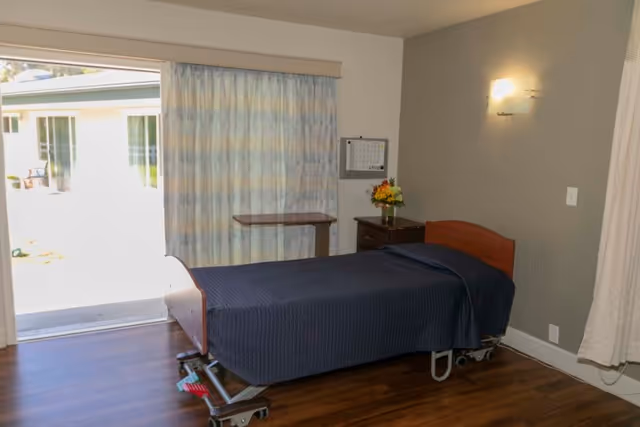 A simple bedroom in a senior living facility with a single bed covered in a dark blue blanket, a small wooden nightstand with a vase of flowers, a wall-mounted light, and a sliding glass door with curtains leading to an outdoor area.