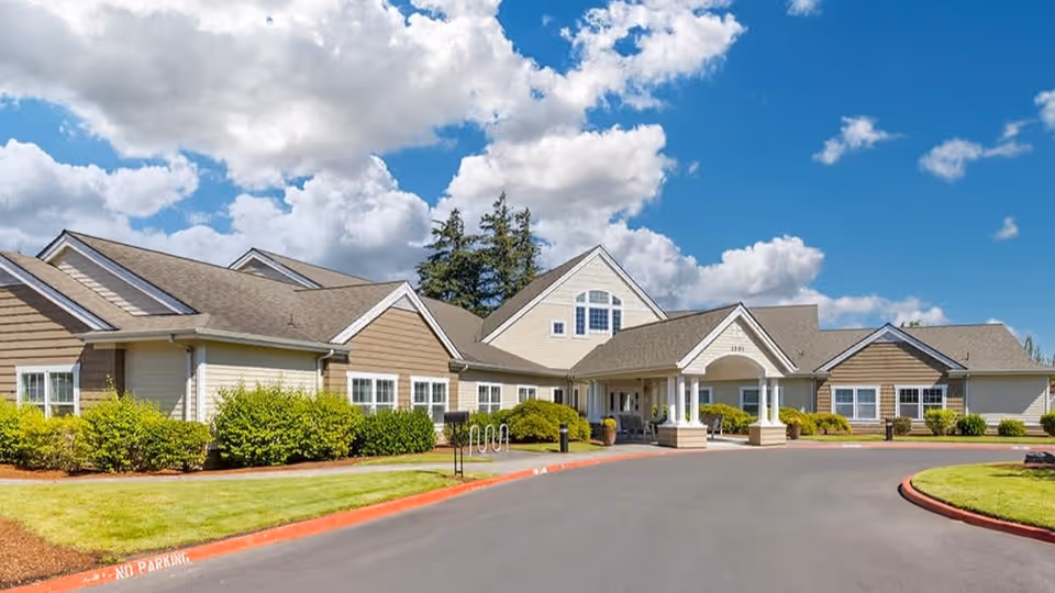 Exterior view of Brookdale Troutdale senior living facility showing a large, single-story building with beige siding, white trim, and a covered entrance. The building is surrounded by well-maintained green bushes and grass under a partly cloudy blue sky.