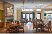 Bright, spacious dining room with round wooden tables set with yellow napkins, chandeliers, large windows, and a stone fireplace.