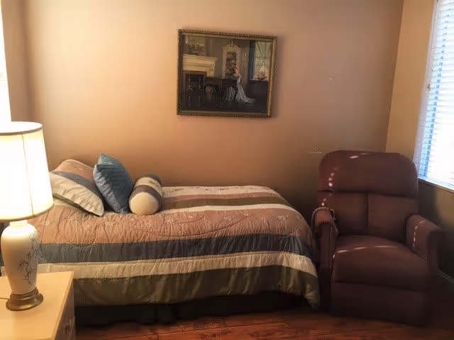 A cozy bedroom with a single bed covered in a striped comforter and several pillows, a bedside table with a lamp, a framed painting on the wall above the bed, and a comfortable recliner chair next to a window with blinds.