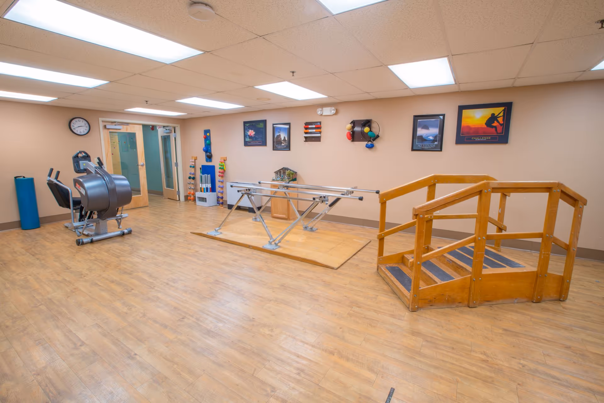 A rehabilitation or physical therapy room with wooden flooring, a set of wooden stairs with handrails for practice, parallel bars for walking exercises, exercise equipment including a recumbent bike, and colorful weights and therapy tools mounted on the wall. The room has beige walls, fluorescent ceiling lights, and a clock above a door leading to another room.