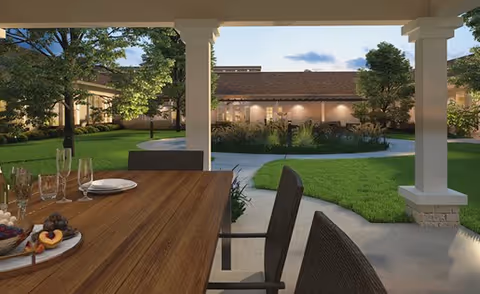 Covered patio with a wooden dining table and chairs overlooking a landscaped courtyard with grass, trees, and a low building.
