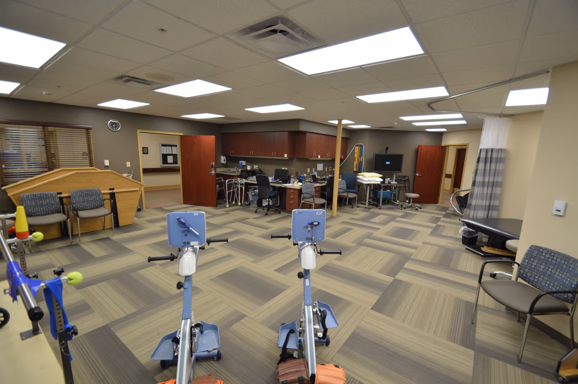 A spacious therapy or rehabilitation room with exercise equipment including two stationary arm bikes in the foreground. The room has patterned carpet flooring, several chairs, a desk with computer equipment, cabinets along the back wall, and a curtained area on the right side. The ceiling has multiple fluorescent lights, and there are open doors leading to other rooms.