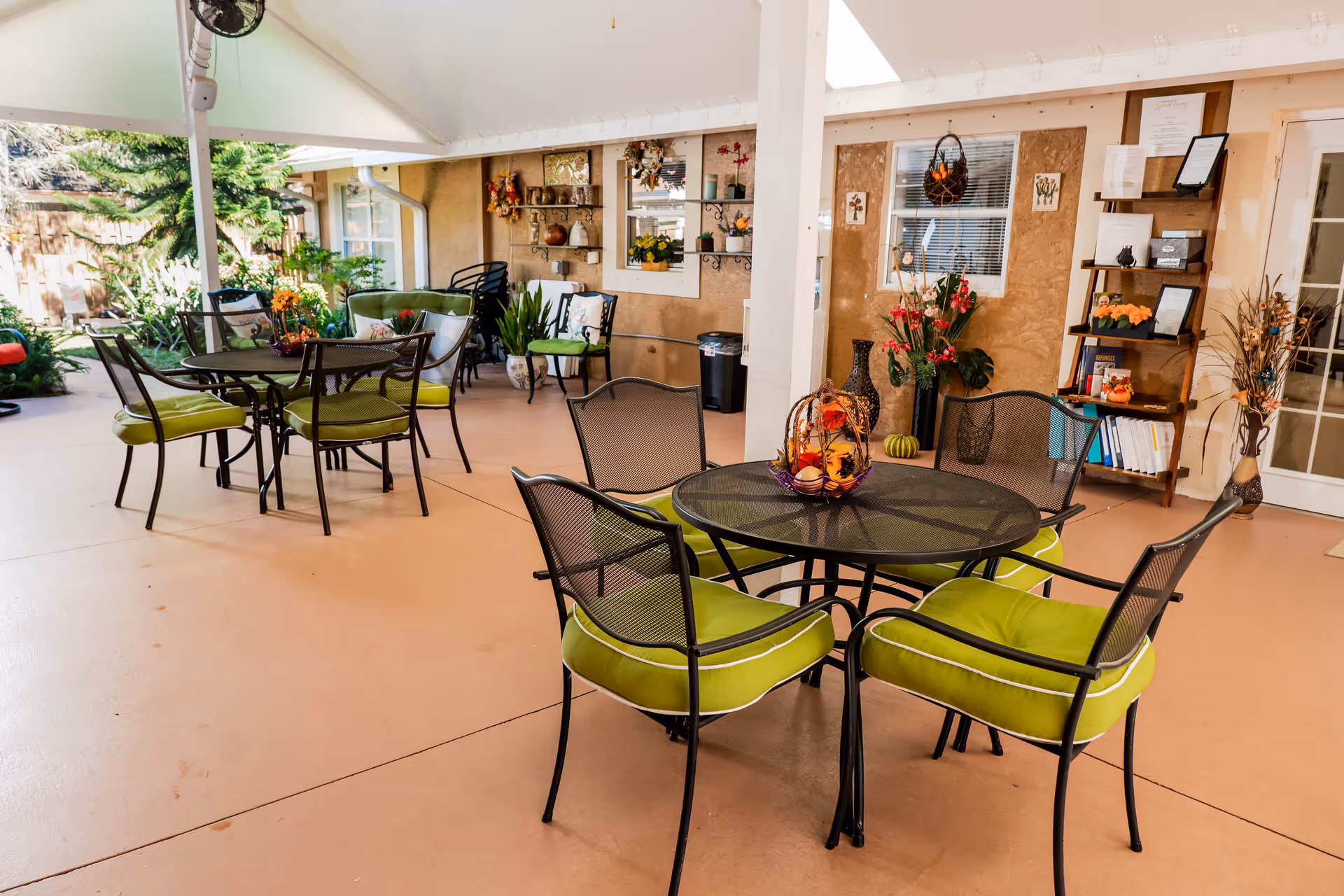 Covered outdoor patio area with several black metal tables and chairs with green cushions. The space is decorated with plants, flowers, and wall shelves holding decorative items. There is a fan mounted on the ceiling and a garden visible in the background.