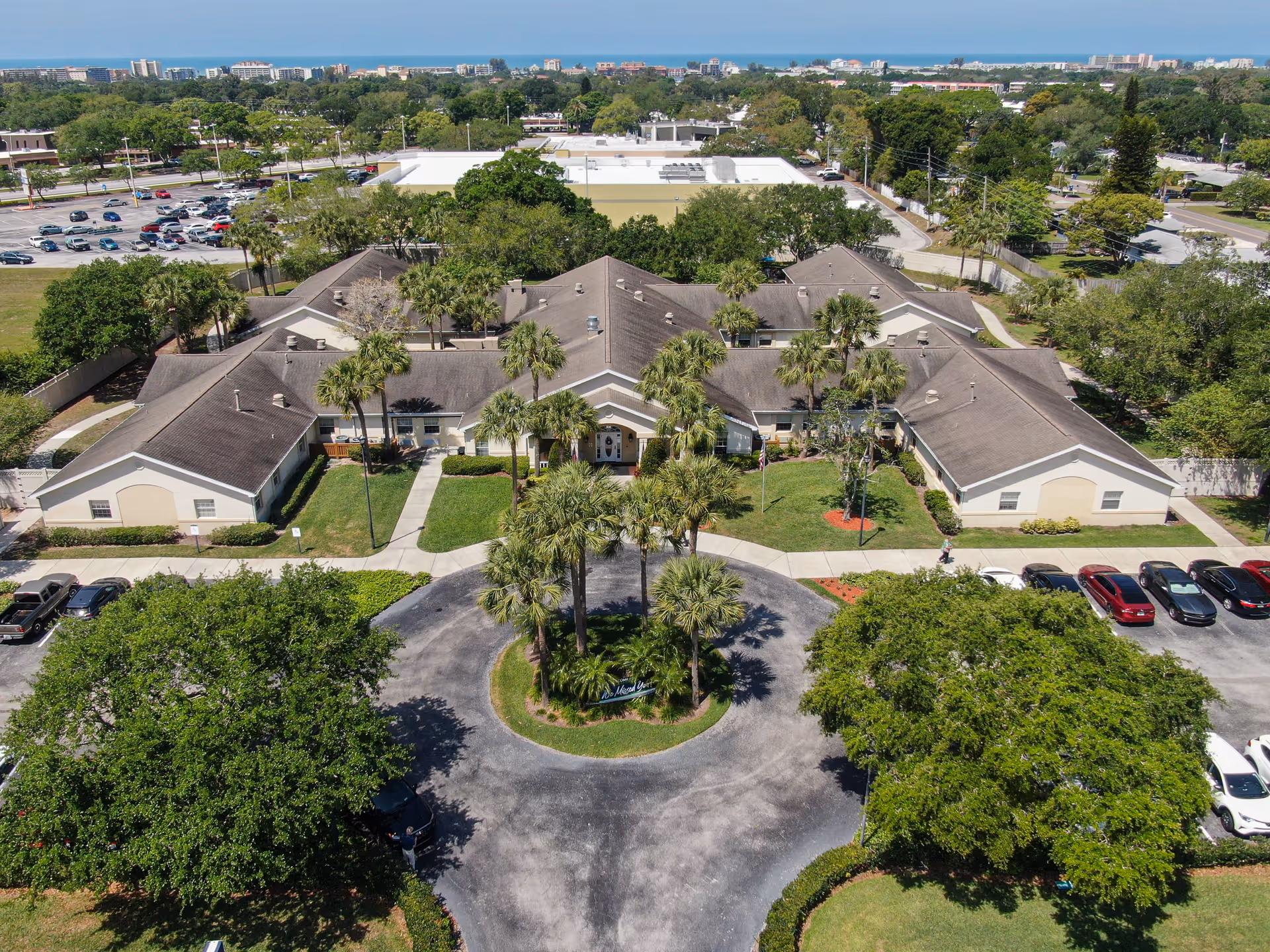 Aerial view of Arden Courts - ProMedica Memory Care Community in Seminole, showing a large single-story building with a circular driveway lined with palm trees at the entrance. The building is surrounded by green lawns, trees, and parking lots with several cars. The background includes more trees, buildings, and a distant view of the ocean under a clear blue sky.