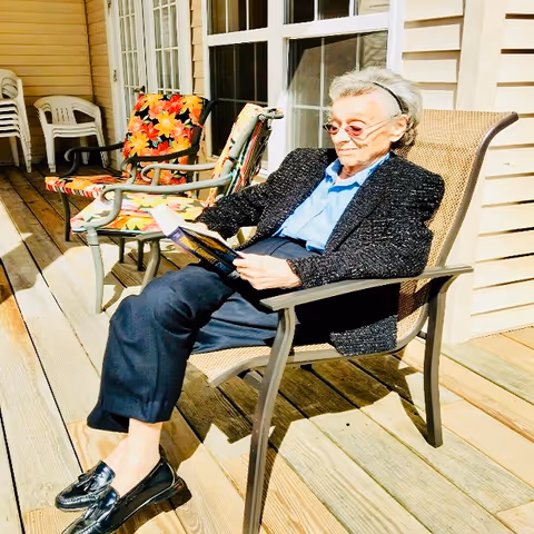 An elderly woman wearing glasses, a black jacket, blue shirt, and dark pants is sitting on a patio chair on a wooden deck, reading a book. Behind her are two chairs with colorful floral cushions and a beige wall with windows.