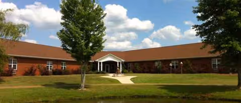 Single-story brick building with a central entrance, bordered by green lawn, trees, and a curved walkway.