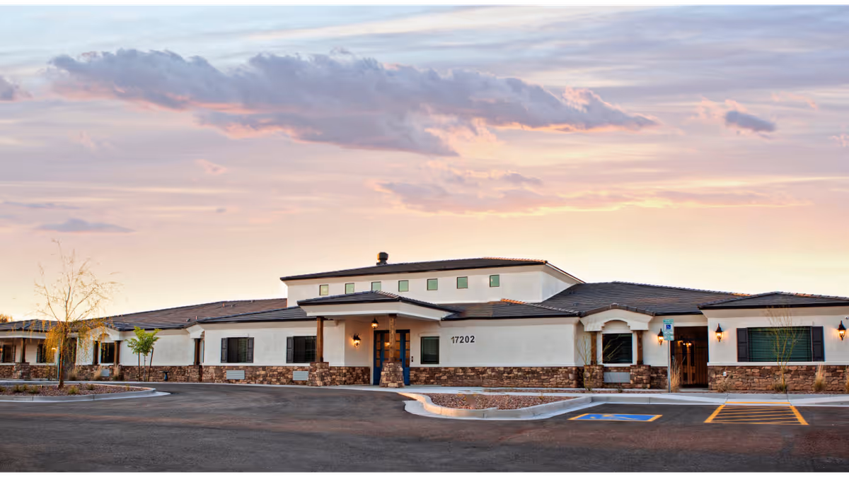Front exterior of a single-story assisted living building with stone trim, an entrance canopy, and a paved driveway at sunset.