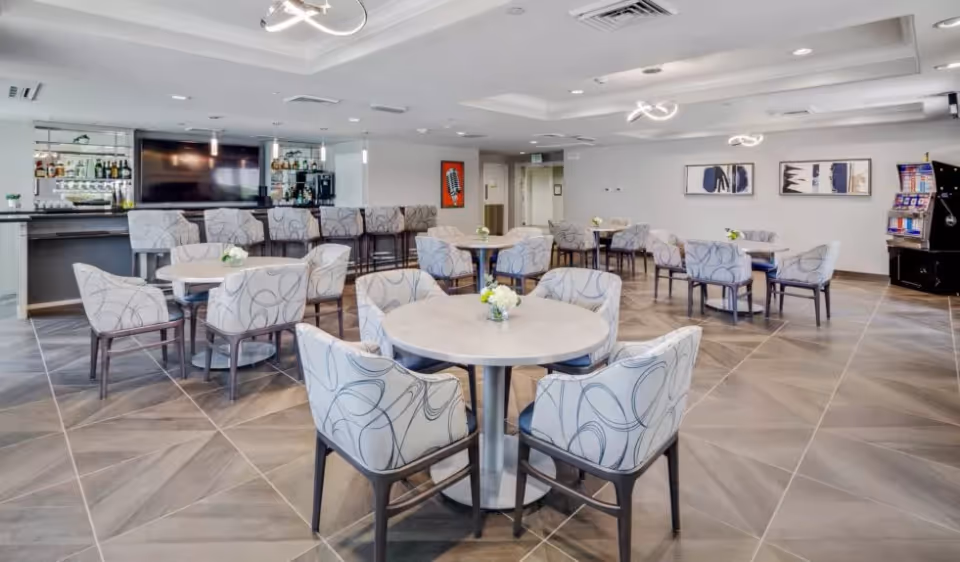 A spacious dining area with multiple round tables surrounded by patterned upholstered chairs. In the background, there is a bar with high stools, a large flat-screen TV mounted on the wall, and shelves stocked with bottles. The room has modern ceiling lights, tiled flooring, and decorative wall art. A slot machine is visible in the far right corner.