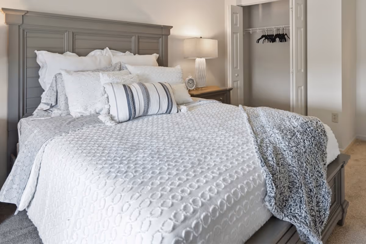 A neatly made bed with a gray wooden headboard, white and patterned pillows, a white textured bedspread, and a gray knitted throw blanket. Next to the bed is a wooden nightstand with a lamp and a small clock. In the background, there is an open closet with several black hangers.