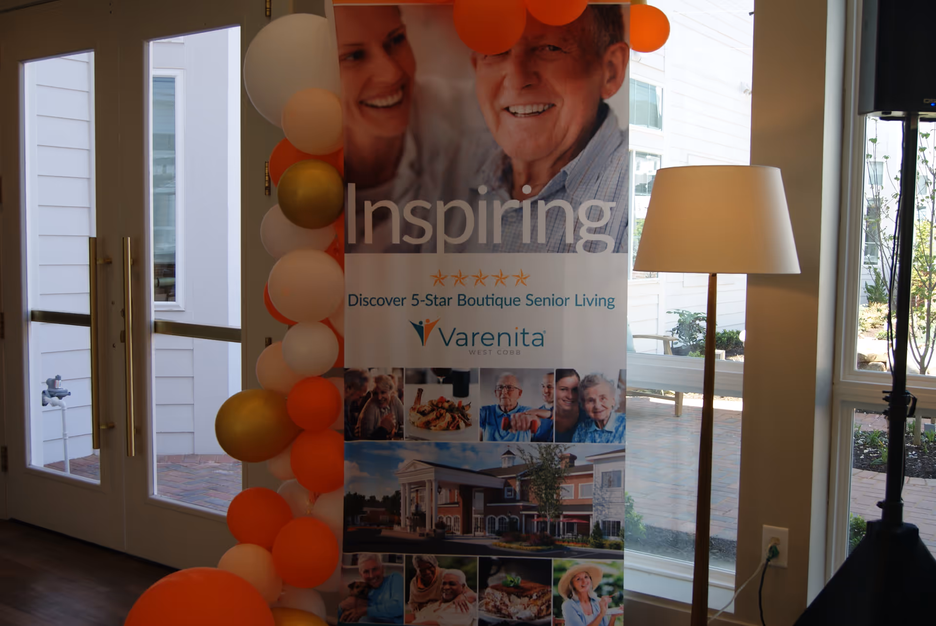 Indoor seating/entry area with a Varenita of West Cobb promotional banner, a column of orange and gold balloons, a floor lamp, and glass doors and windows.