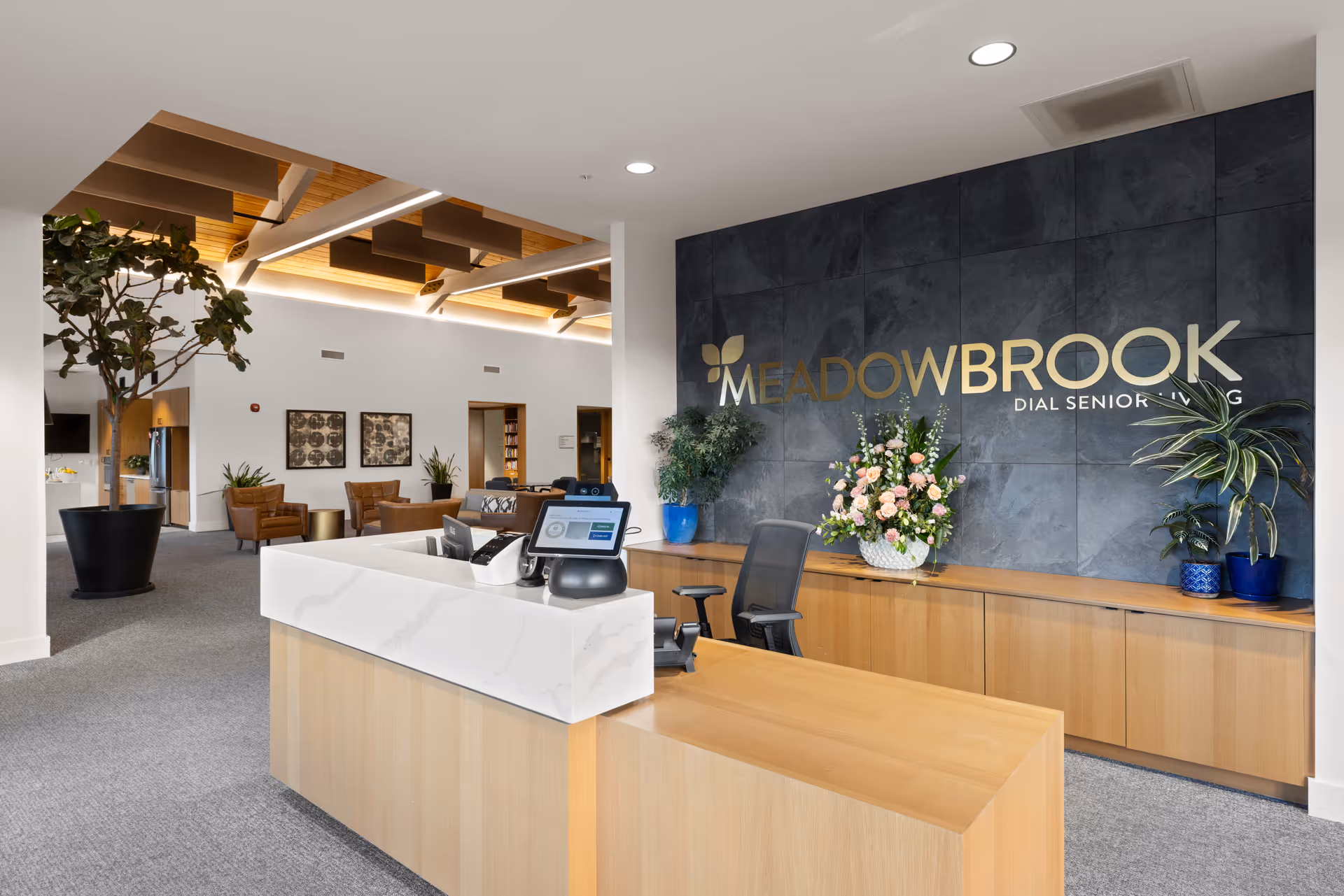 Reception area of Meadowbrook Senior Living - Dial Senior Living featuring a modern wooden reception desk with a computer and phone, a dark tiled wall with the facility's name in gold letters, several potted plants, and a seating area with chairs and framed artwork in the background.