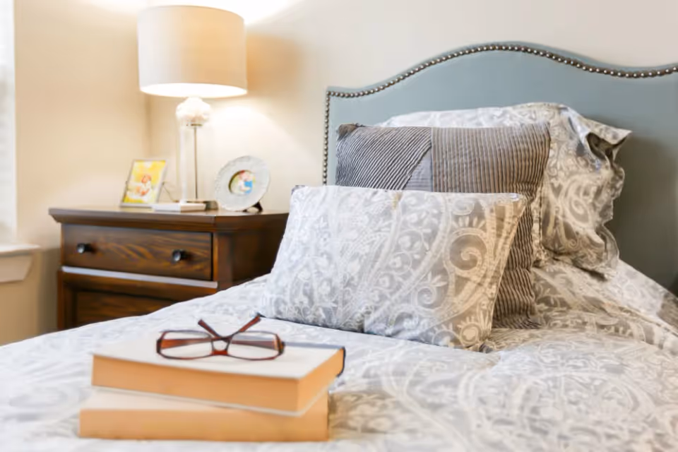 A neatly made bed with patterned gray and white bedding and multiple pillows, a pair of eyeglasses resting on two stacked books on the bed. Next to the bed is a wooden nightstand with a lamp, a framed photo, and a decorative plate.