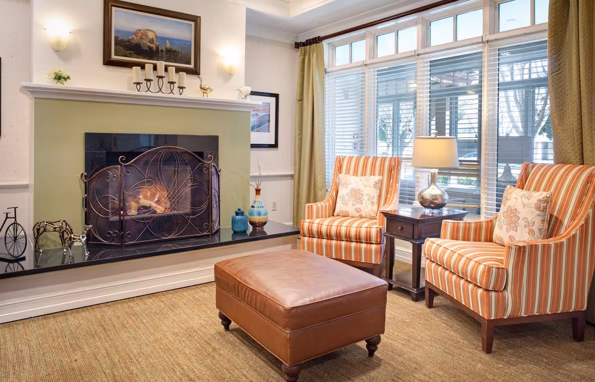 Cozy living room with a lit fireplace, two striped armchairs, a leather ottoman, and large windows with blinds.