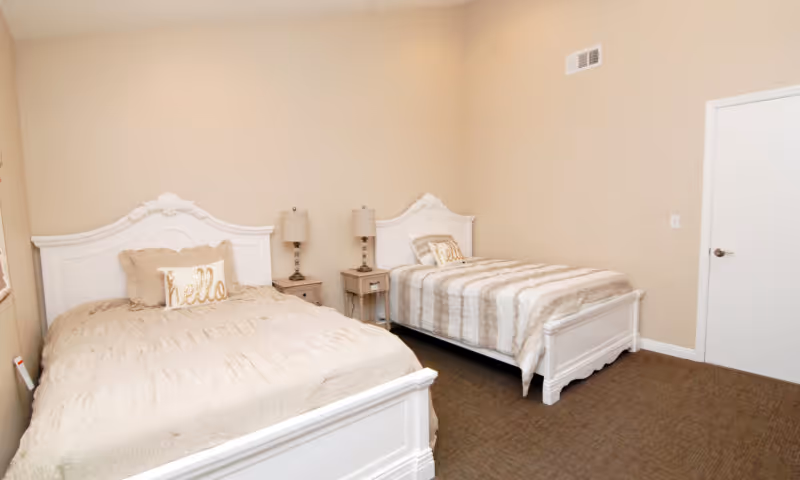 A neutral-toned bedroom with two white-framed beds, a shared nightstand and lamps between them.
