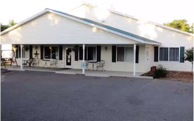 Single-story building with white siding and a green roof, featuring a covered porch with several small tables and chairs, a swing on the left side, and windows with shutters. The building is surrounded by a paved area and some landscaping with bushes and trees.